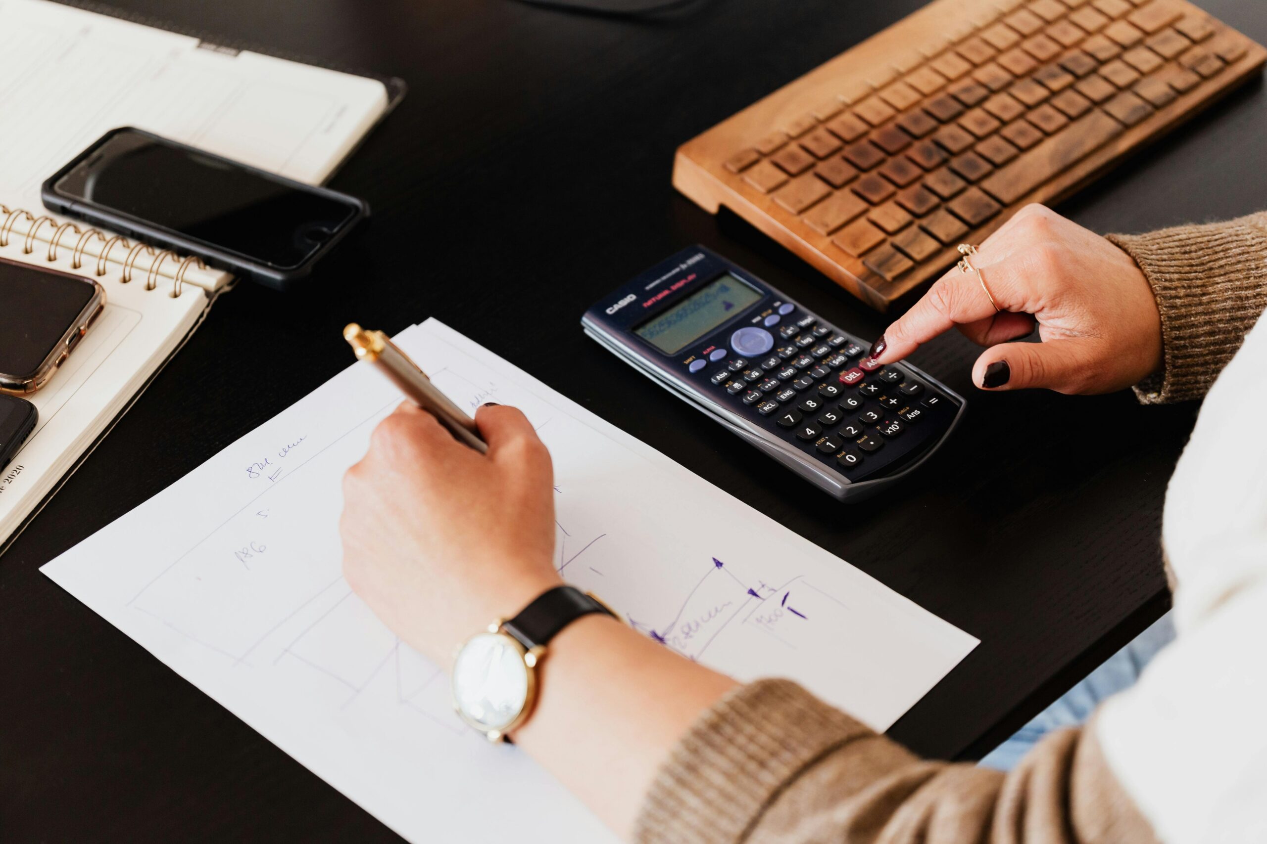 From above crop unrecognizable female accountant wearing stylish watches using calculator and writing down information on paper while working at desk with gadgets